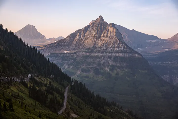 Mountain peaks at sunrise above a thin road cut into the slope on Going-to-the-Sun Road in Glacier National Park